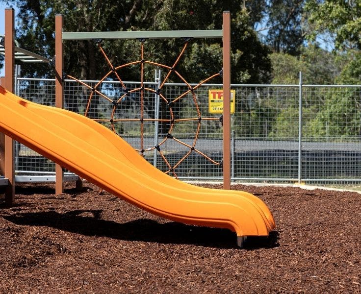 The double slide of the new playground and the spider rope climb are visible, surrounded by a fence as the playground is still under construction.