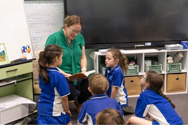 Principal reading a story in the classroom to 5 students, students are looking on and enjoying the story.