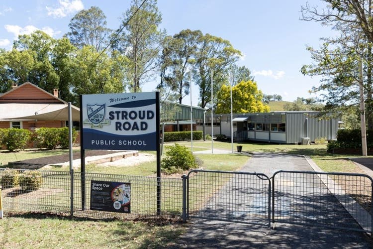 Outside view of the front of the school from the front gate. The school sign is in the foreground and in the background the classroom, cola and office building a shown, surrounded by trees.
