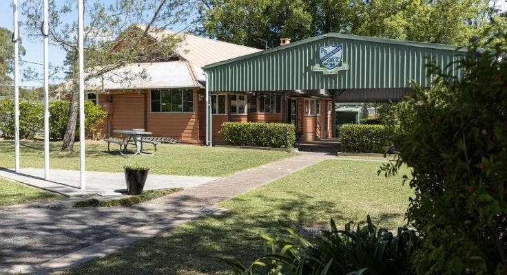 View of the Cola and the classroom. The flag poles and gardens are in the foreground and the school logo sits at the top of the cola.