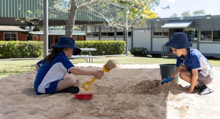 Two students playing together in the sandpit, both have shovels and the office building is in the background.